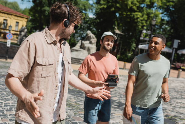guía turístico en auriculares gesticulando cerca de turistas multiétnicos caminando con el mapa y la cámara vintage en el descenso de Andrews en Kiev - Foto, Imagen