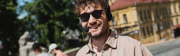 portrait of carefree tour guide in sunglasses and headset smiling at camera on blurred Andrews descent in Kyiv, banner - Photo, Image