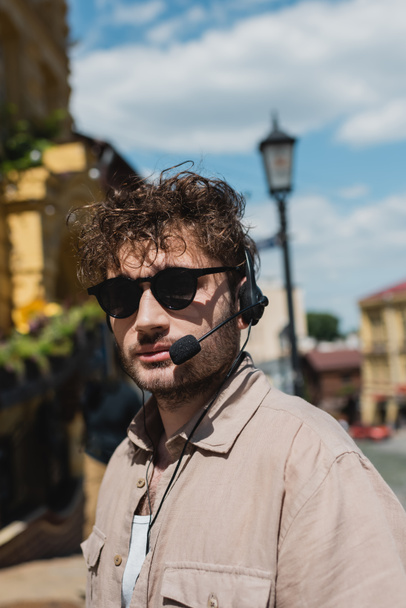 портрет молодого экскурсовода в солнцезащитных очках и наушниках, смотрящего в камеру на размытом спуске Эндрюса в Киеве - Фото, изображение