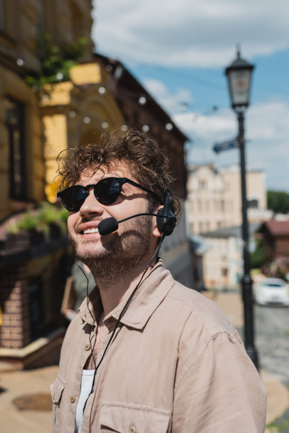 молодой и кудрявый экскурсовод в солнцезащитных очках и наушниках, улыбающийся на размытом спуске Эндрюса в Киеве - Фото, изображение