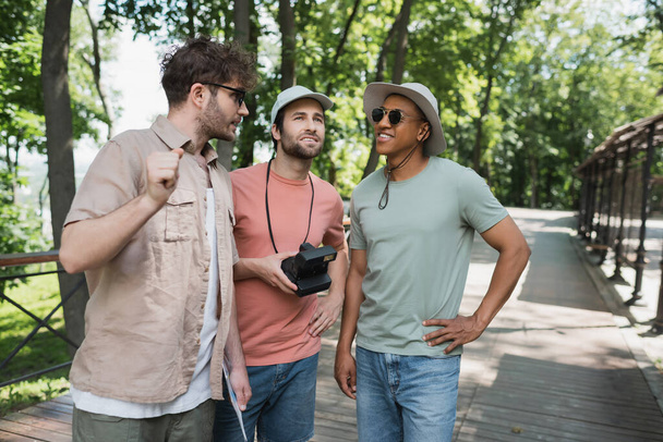 bebaarde toerist met vintage camera op zoek weg in de buurt van multi-etnische mannen tijdens excursie in het zomerpark - Foto, afbeelding