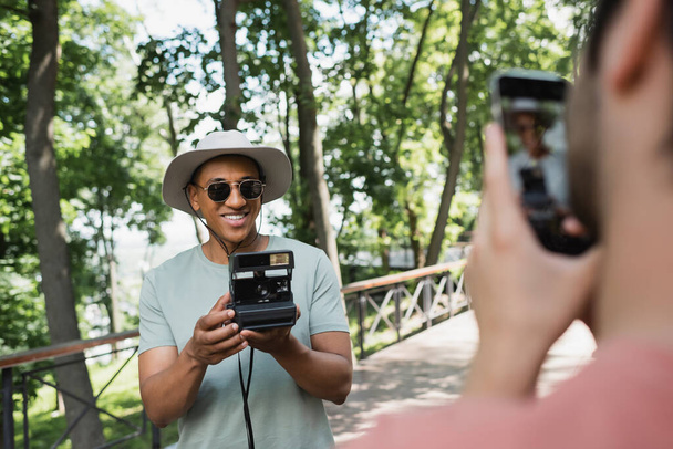 blurred man with smartphone taking photo of carefree african american tourist in sunglasses posing with vintage camera in park - Photo, Image