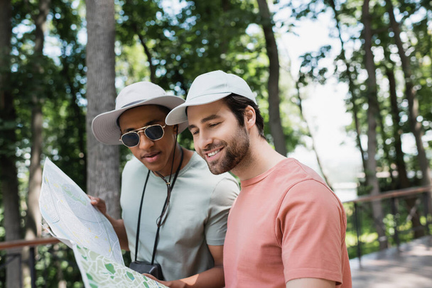 pozitivní multietničtí turisté ve slunečních čepicích při pohledu na cestovní mapu v rozmazaném parku - Fotografie, Obrázek