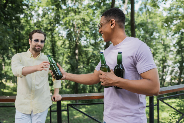 carefree african american man giving bottle of beer to friend in city park - Foto, Imagen