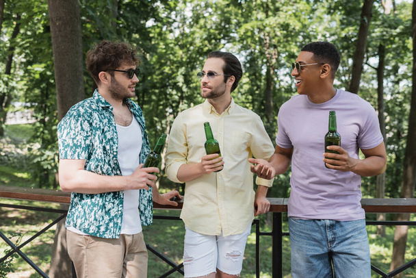 fröhlicher afrikanisch-amerikanischer Mann im Gespräch mit trendigen Freunden, die mit Bier am Zaun im Stadtpark stehen - Foto, Bild