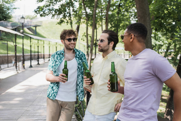 beztroskie międzyrasowe przyjaciele w stylowy letni strój trzymając butelki piwa i rozmowy w miejskim parku - Zdjęcie, obraz