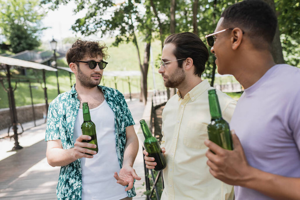 стильный мужчина в солнечных очках держит пиво и жестикулирует, разговаривая с многонациональными друзьями в городском парке в летний день - Фото, изображение