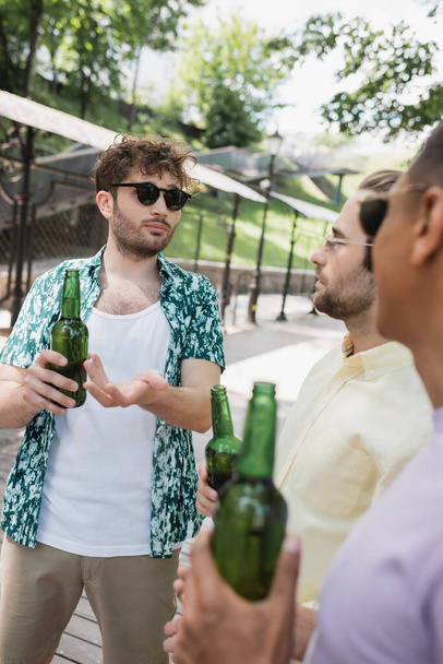 młody i modny mężczyzna w okularach przeciwsłonecznych wskazujący na butelkę piwa w pobliżu rozmazanych przyjaciół międzyrasowych w parku miejskim - Zdjęcie, obraz