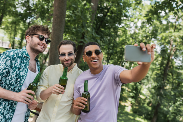 african american man holding beer and taking selfie with carefree friends in summer park - Photo, Image