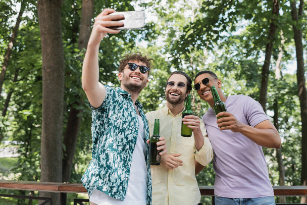 счастливый и модный мужчина в солнечных очках делает селфи на смартфоне с межрасовыми друзьями с пивом в зеленом парке - Фото, изображение
