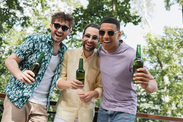 amigos multiétnicos despreocupados en traje elegante y gafas de sol sosteniendo cerveza y riéndose de la cámara en el parque - Foto, imagen