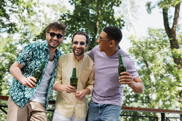 radosnych i modnych przyjaciół w okularach przeciwsłonecznych trzymających piwo i śmiejących się w zielonym parku - Zdjęcie, obraz