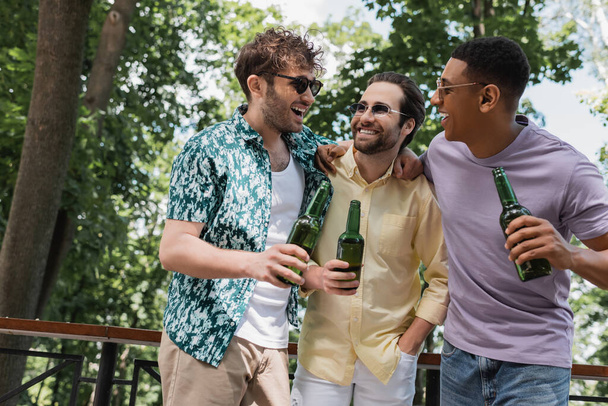 amigos despreocupados multiculturales en traje de verano elegante abrazando mientras sostiene la cerveza en el parque de la ciudad - Foto, imagen