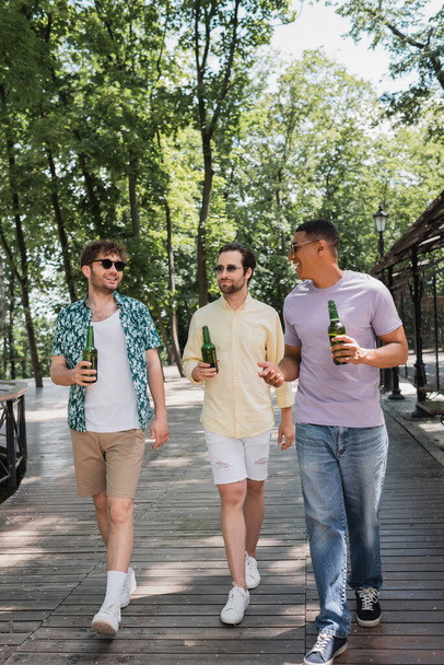 夏の公園を歩きながらビール瓶を持った流行の服を着た幸せな多文化の友人の完全な長さ - 写真・画像