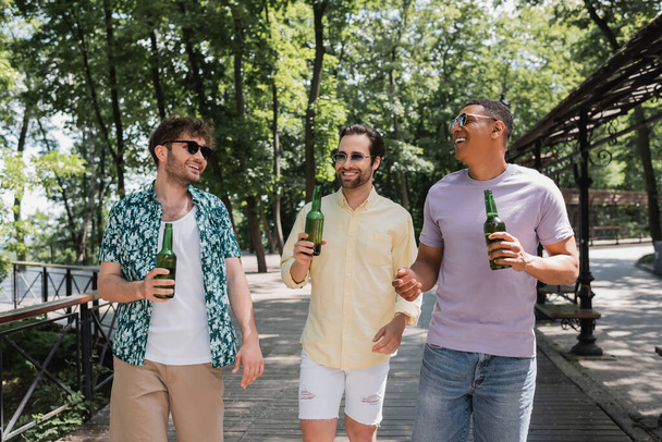 carefree and stylish multiethnic friends holding beer while walking in urban park on summer day - Foto, Imagen