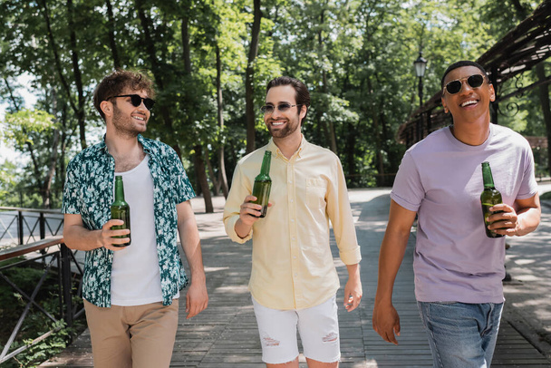 amigos multiétnicos alegres y de moda sosteniendo cerveza fresca durante el paseo en el parque de la ciudad en el día de verano - Foto, imagen