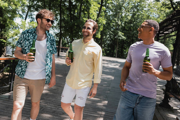 amigos despreocupados multiculturales en traje de verano de moda y gafas de sol caminando con cerveza en el parque urbano - Foto, Imagen