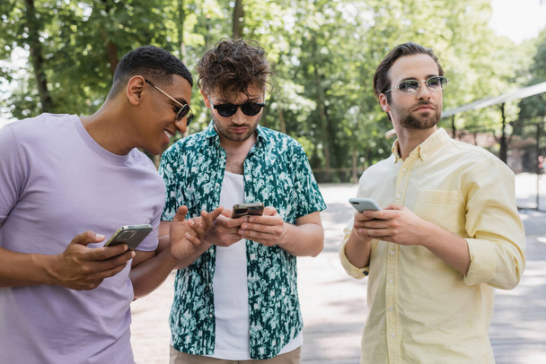 joyeux homme afro-américain pointant du doigt près des amis à la mode en utilisant des téléphones cellulaires dans le parc d'été - Photo, image
