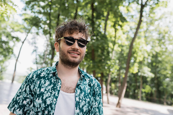 Uśmiechnięty mężczyzna w luźnych ubraniach i okularach słonecznych w letnim parku  - Zdjęcie, obraz