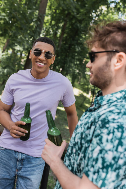 Uśmiechnięty Afroamerykanin w okularach przeciwsłonecznych trzymający piwo w pobliżu rozmazanego przyjaciela w letnim parku  - Zdjęcie, obraz