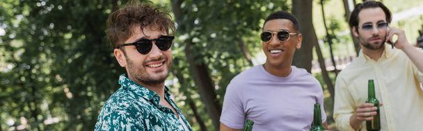 Joven con gafas de sol de pie cerca de amigos interracial con cerveza en el parque de verano, pancarta  - Foto, Imagen