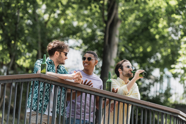 Amigos multiétnicos positivos en gafas de sol hablando cerca de la cerveza en barandilla en el parque de verano  - Foto, imagen
