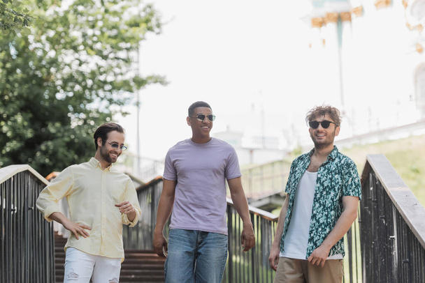 Positive interracial men in sunglasses walking on stairs in summer park  - Foto, Imagem