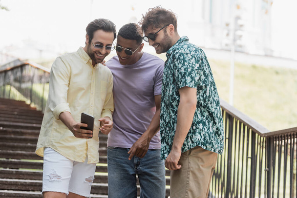 Mladý muž ukazuje mobilní telefon multietnické přátele ve slunečních brýlích na schodech v parku  - Fotografie, Obrázek