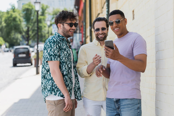 Carefree interracial friends in sunglasses using mobile phone on street in Kyiv  - 写真・画像