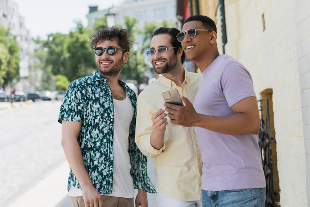 Usmívající se mezirasoví přátelé ve slunečních brýlích drží mobilní telefon a dívá se jinam na ulici v Kyjevě  - Fotografie, Obrázek