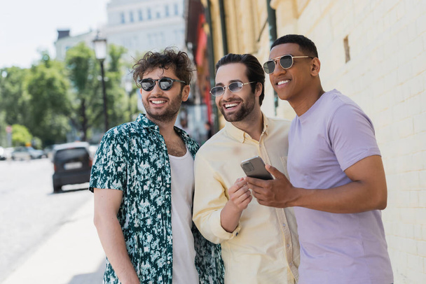 Uomini multietnici sorridenti in occhiali da sole che tengono il cellulare e distolgono lo sguardo sulla strada a Kiev  - Foto, immagini