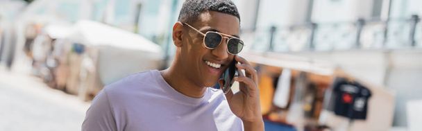 Homme afro-américain positif en lunettes de soleil parlant sur smartphone dans la rue urbaine, bannière  - Photo, image