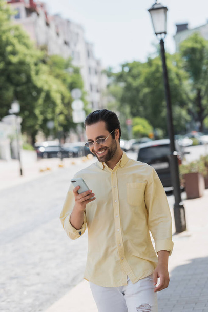 Cheerful man in shirt and sunglasses using smartphone while walking on urban street  - Foto, Bild