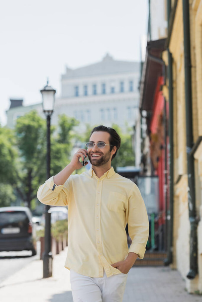 Happy brunette mies aurinkolasit puhuu älypuhelimella kävellessään kadulla Kiovassa  - Valokuva, kuva