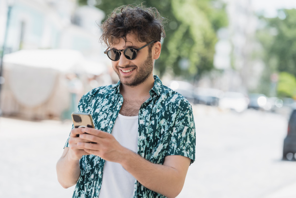 Positive brunette man in sunglasses using mobile phone on blurred urban street  - 写真・画像