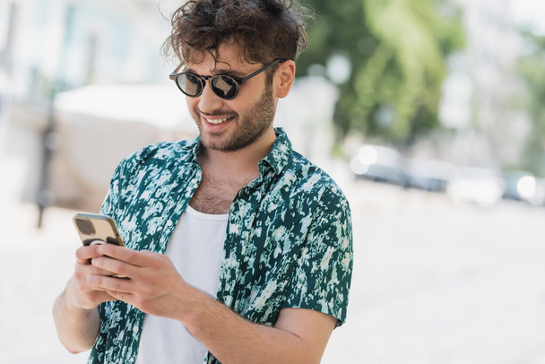 Carefree man in sunglasses using smartphone on blurred urban street in summer  - Photo, Image