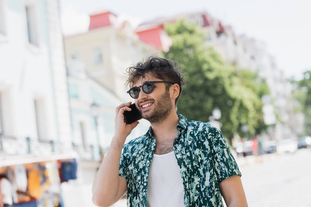 Positive young man in sunglasses talking on smartphone while walking on urban street  - Fotoğraf, Görsel