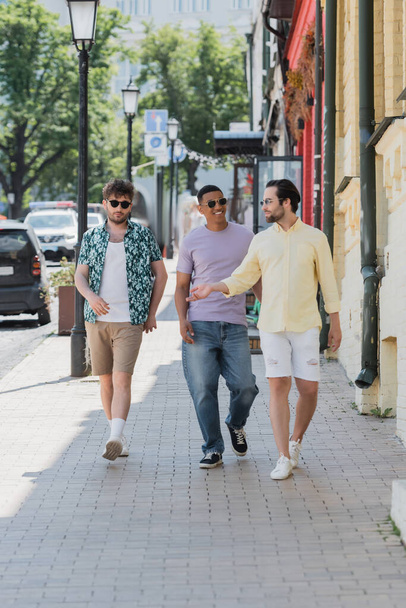 Smiling interracial friends in sunglasses walking on Andrews descent in Kyiv  - Photo, Image