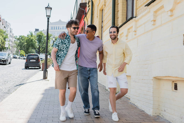 Sorrindo amigos inter-raciais em óculos de sol andando na calçada na descida Andrews em Kiev  - Foto, Imagem