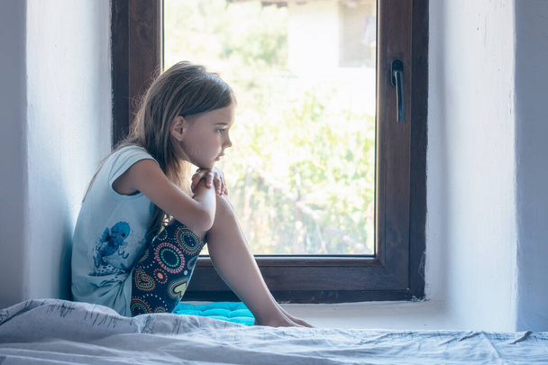 Vintage portrait of a beautiful little girl with bare feet and pensive gaze sitting near a window inside a room	 - Foto, afbeelding