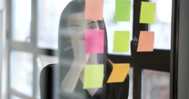 Manager vrouw schrijven herinnering op veelkleurige stickers op glas in kantoor 4k film slow motion. Organisatie van de arbeid in het bedrijfsleven - Video