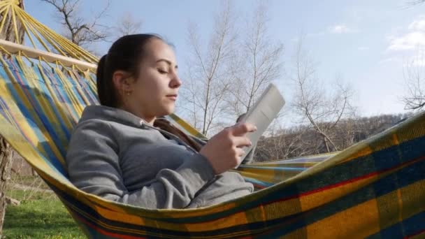La chica está descansando en una hamaca en la naturaleza con una tableta en sus manos. - Imágenes, Vídeo
