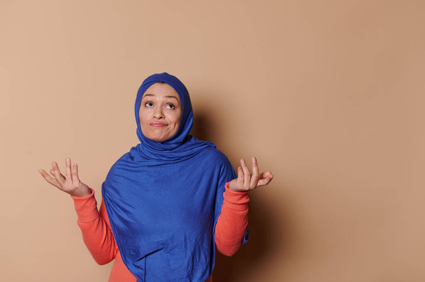 Thoughtful Arabic Muslim woman in a blue hijab, looking up with uncertainty, holding her hands palms up, standing isolated on a beige background. People. Concepts. Lifestyle - Photo, Image