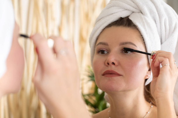 An attractive young woman paints her eyelashes with mascara in front of the bathroom mirror. Make-up. - Foto, immagini