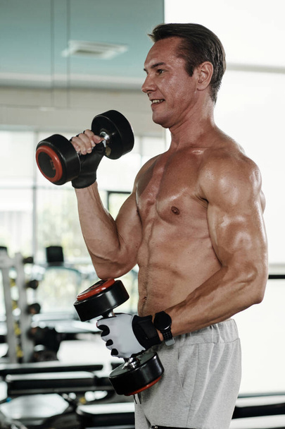 Smiling fit mature man doing biceps exercise with dumbbells - Foto, Bild
