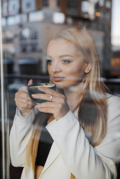 Blond stijlvolle vrouw bij oversized witte jas drinken aroma koffie en ontspannen in cafe - Foto, afbeelding
