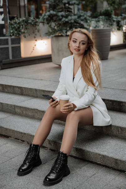 Blond stylish woman at oversized white jacket using phone and drinking aroma coffee on the modern city background - Zdjęcie, obraz