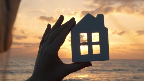 Primer plano de una mujer sosteniendo una silueta de una casa por detrás. La escena tiene lugar en la orilla del océano durante la puesta del sol, con la luz del sol pasando a través de la ventana de la casa. El video transmite - Imágenes, Vídeo