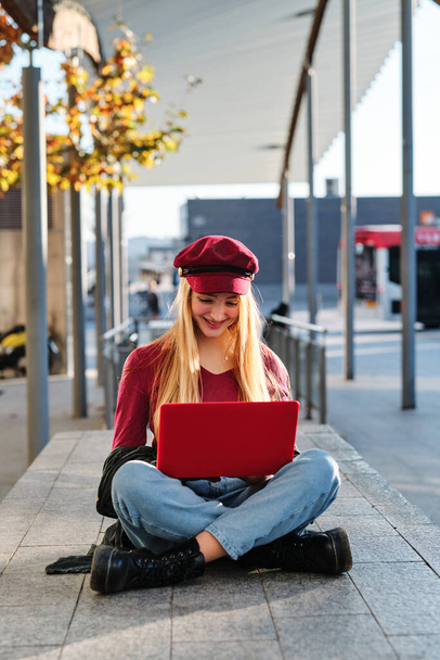 Vertical photo of a blonde and fashionable woman smiling while using a laptop sitting on a city square - 写真・画像
