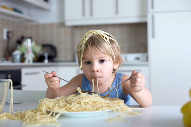 Little blond boy, toddler child, eating spaghetti for lunch and making a mess at home in kitchen - Zdjęcie, obraz